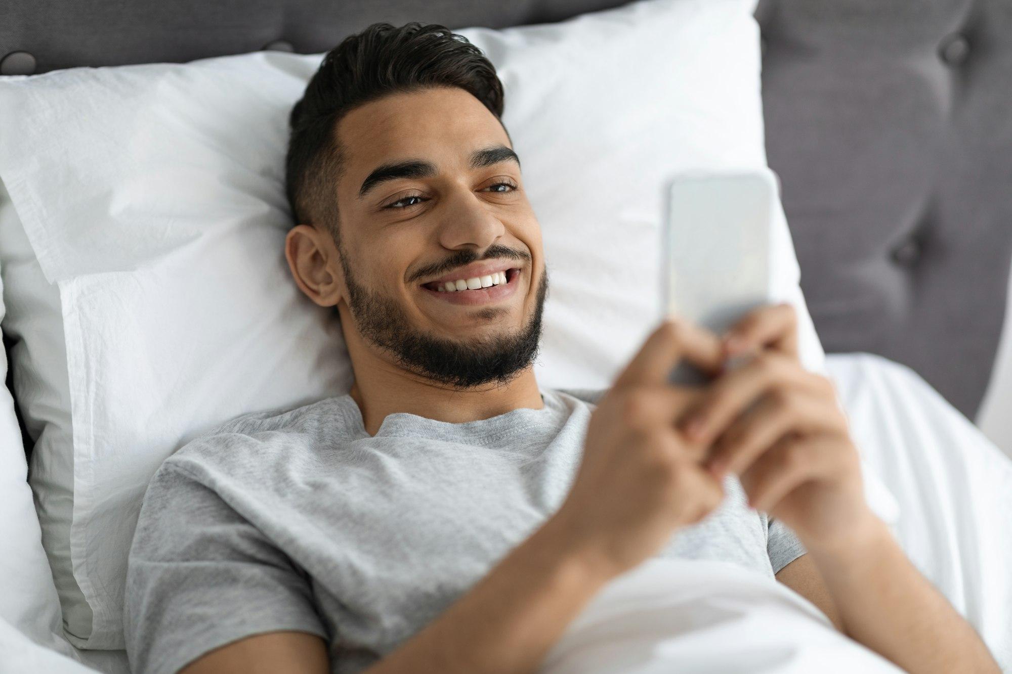 Mobile Gaming. Cheerful Young Arab Man Using Smartphone While Lying In Bed