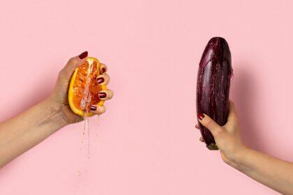 Female hand with half of juicy orange and eggplant on pink background. Sex concept.