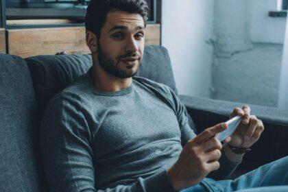 Handsome man looking at camera while watching porn on smartphone on couch