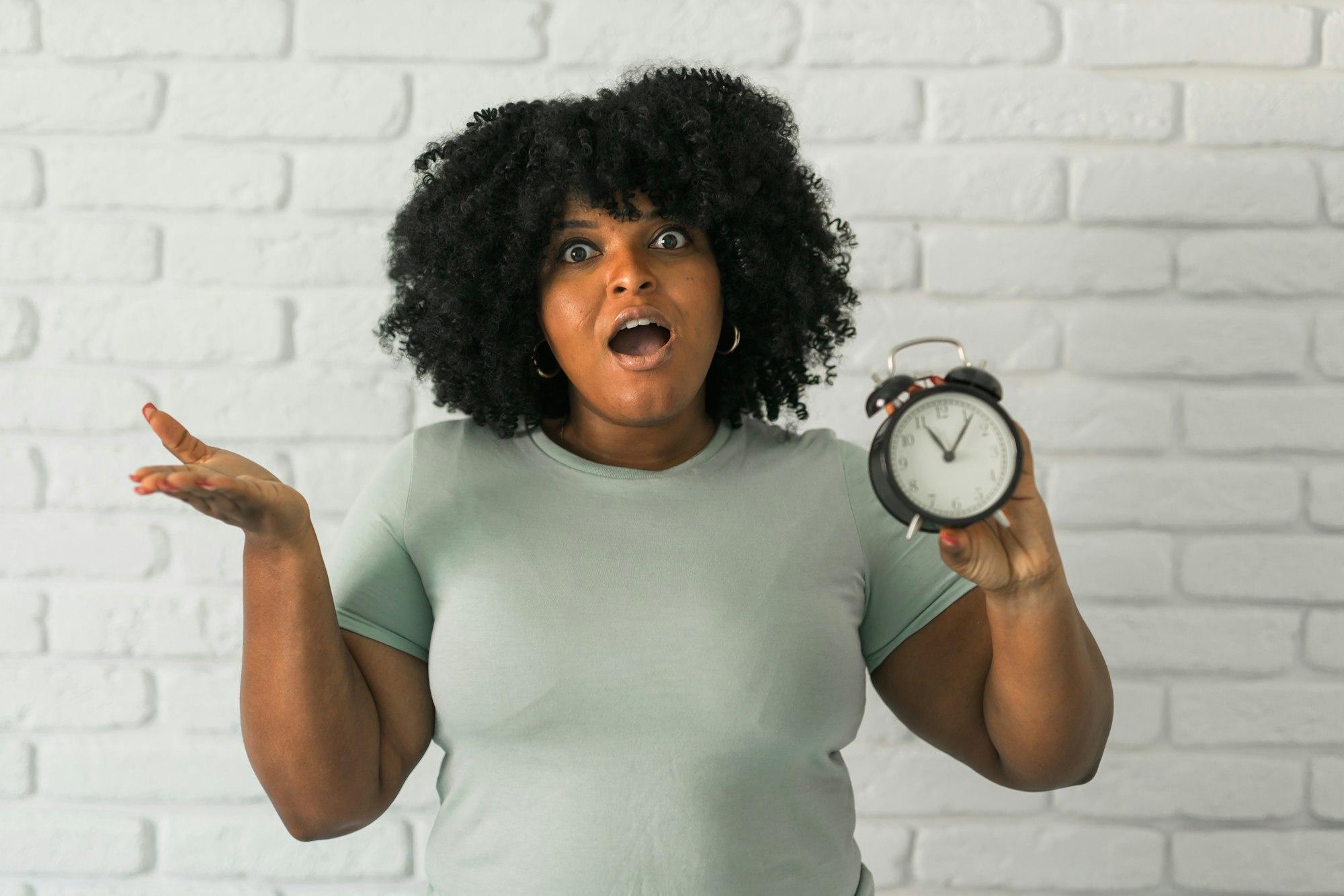 Surprised african american woman holding alarm clock in one hand on brick wall background. Deadline