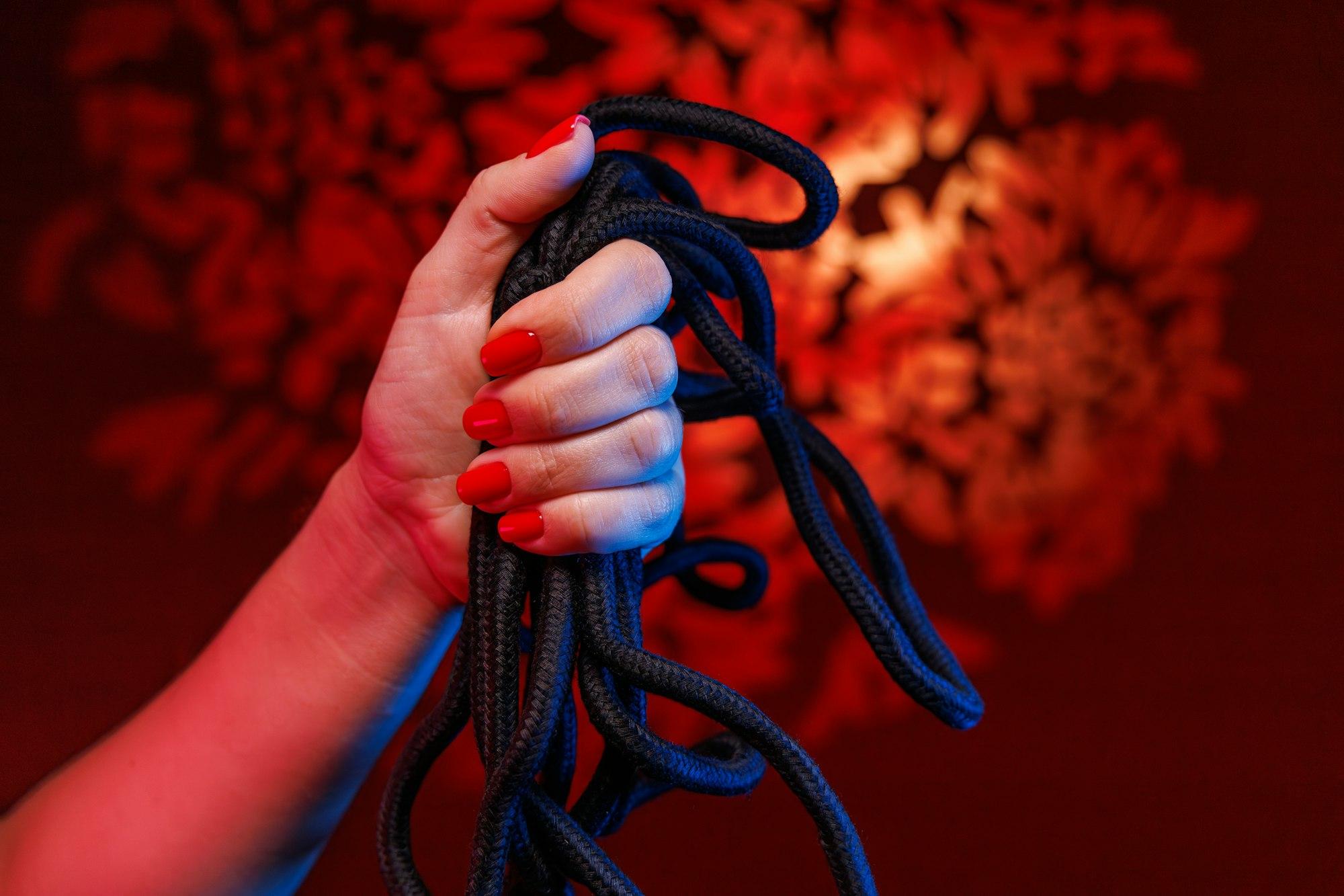 A delicate hand with a sexy red manicure holds a Shibari rope emphasizing the beauty of bondage art