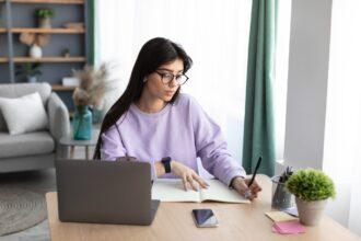 Woman sitting at desk, using computer and writing in notebook