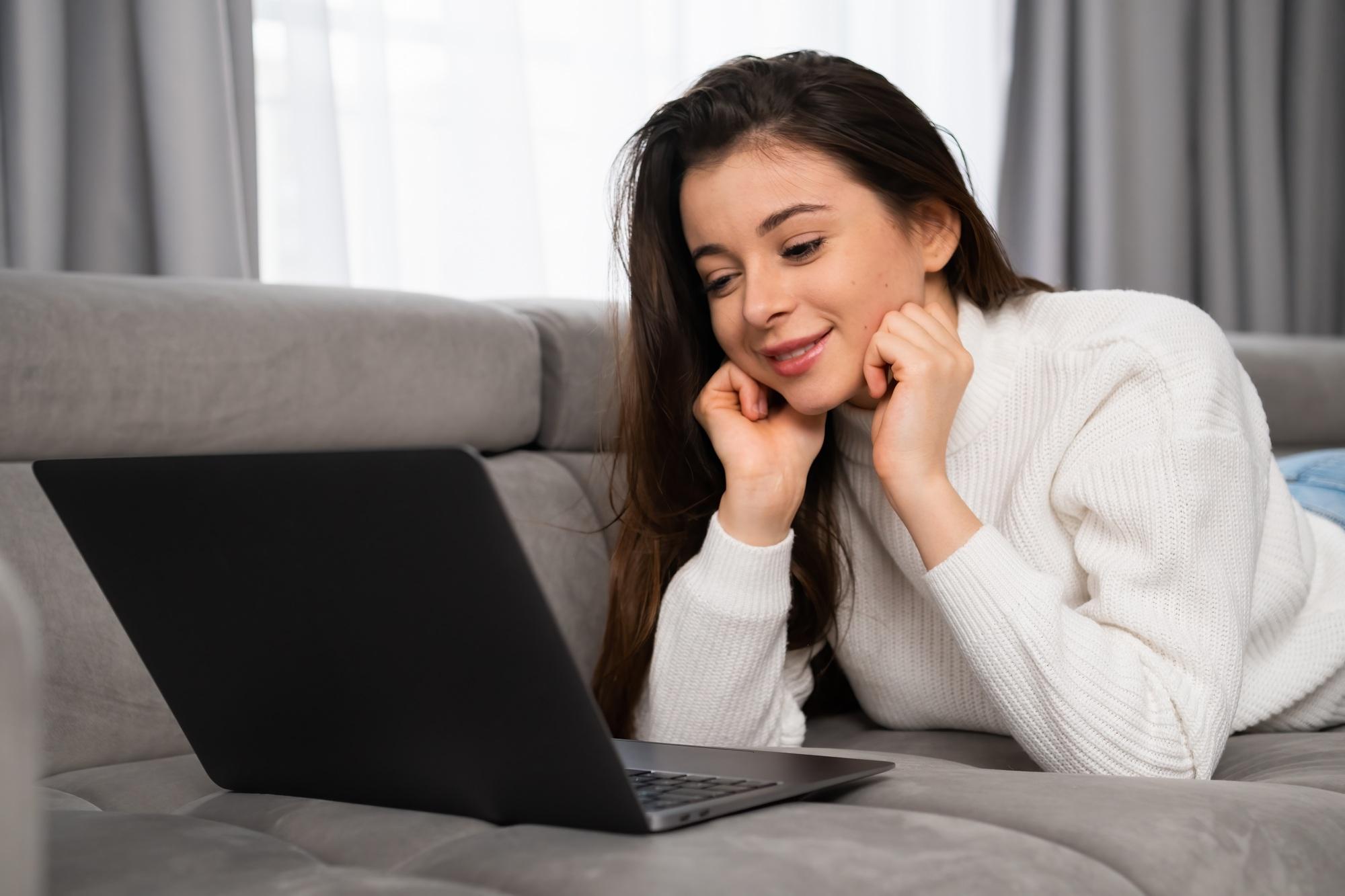 A smiling girl has online dating via laptop, lying on the sofa at the apartment