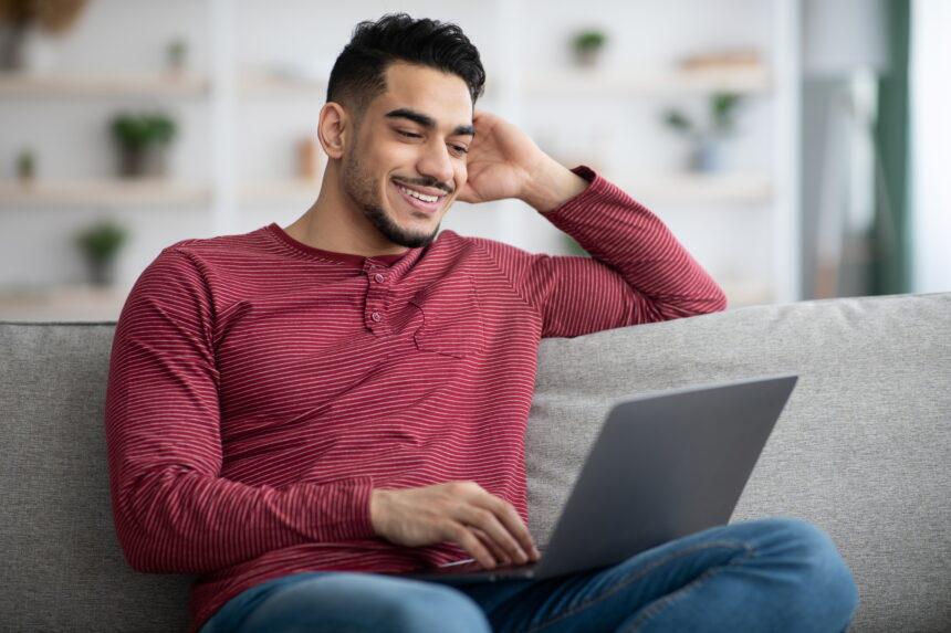 Happy arab guy using laptop, chatting on dating website