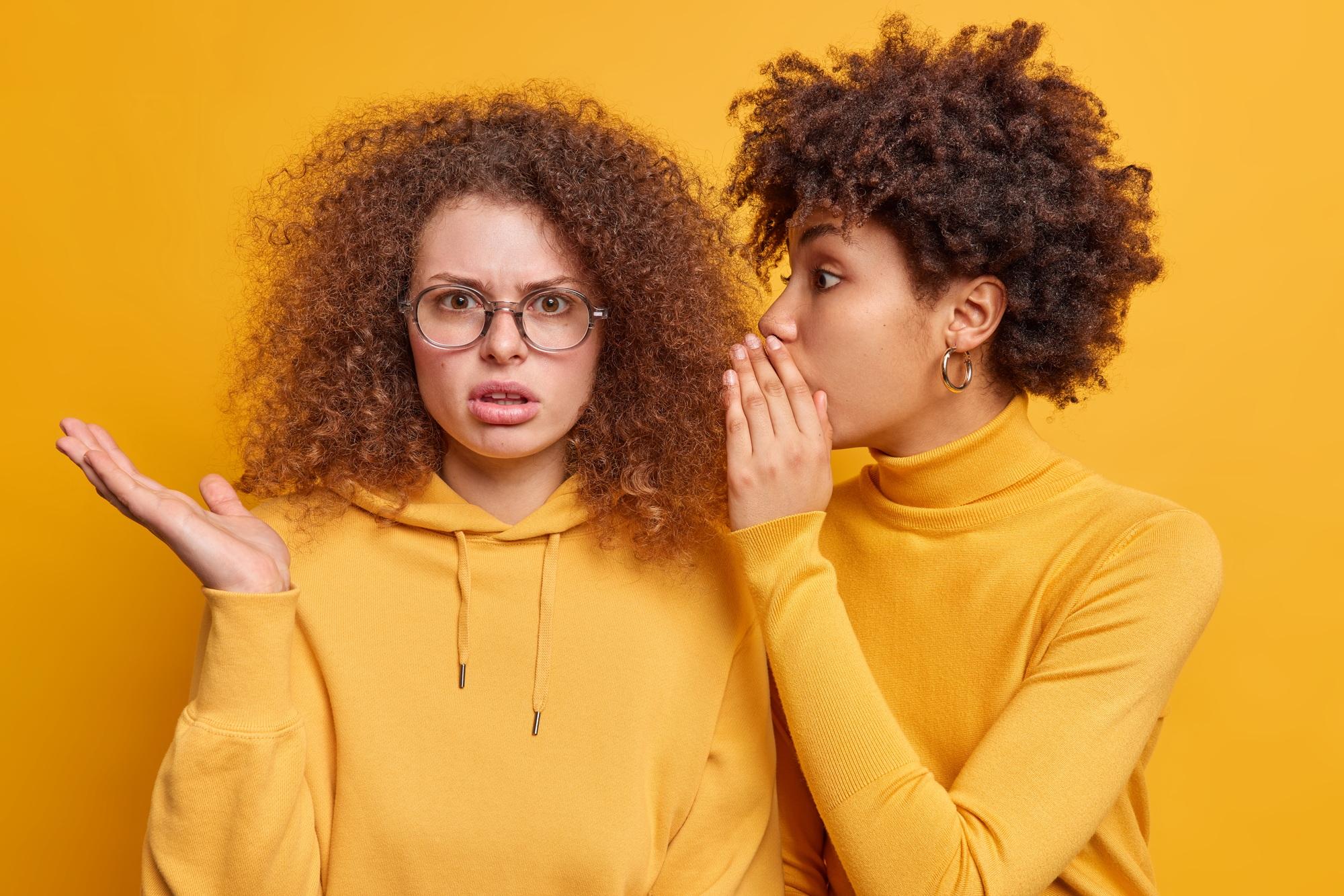 Surprised Afro American woman whispers secret to friend tells secretly something on ear share gossip